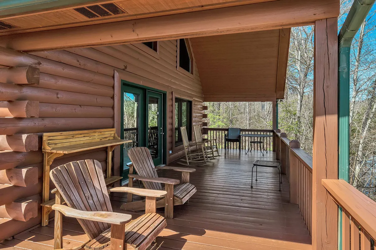 A spacious wooden deck overlooking the surrounding forest, furnished with comfortable chairs and bordered by large windows. The deck is designed for relaxing, gathering, and enjoying the tranquil natural setting.