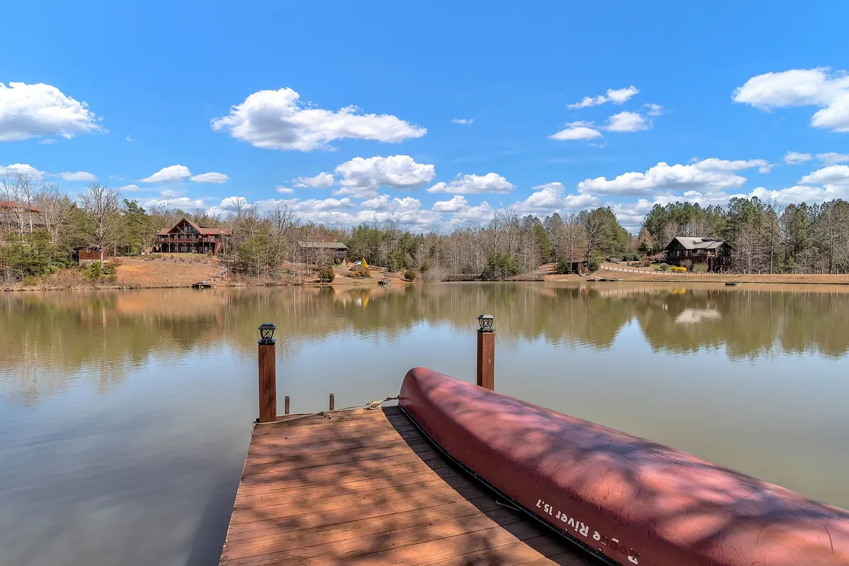 A peaceful lake view at sunset, with a wooden dock jutting into the calm water. A canoe is tied nearby, and the shoreline is framed by leafy trees. Distant cabins dot the horizon, and the tranquil scene invites both relaxation and gentle adventure.