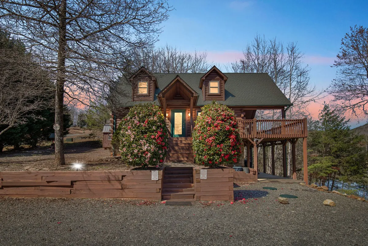 A welcoming exterior view of the cabin's wooden front porch, framed by vibrant flower bushes and a stone pathway, with a gabled roof overhead. The scene is tranquil, inviting, and exudes neighborly charm, suggesting a well-maintained, peaceful community setting.