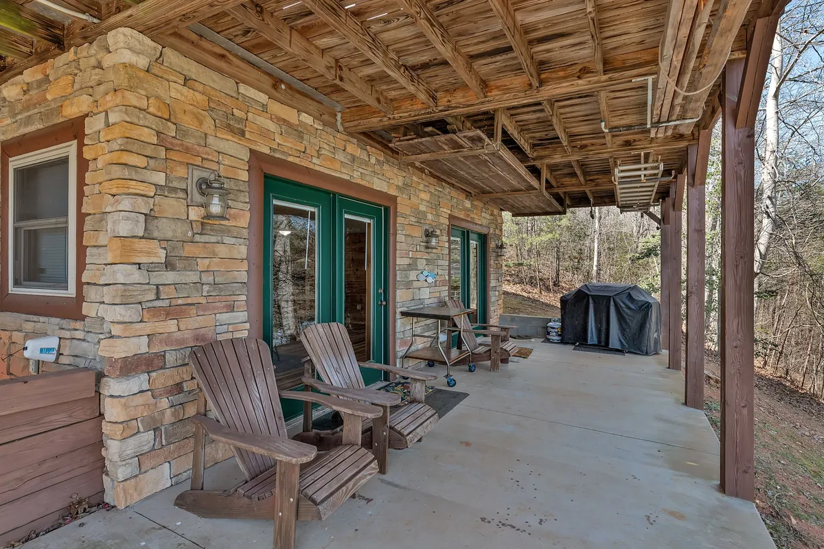 A pair of wooden Adirondack chairs sit atop a stone and wood patio, surrounded by greenery and facing out toward nature. An outdoor grill is nearby, suggesting a space for evening gatherings, relaxation, and stargazing beneath open skies.