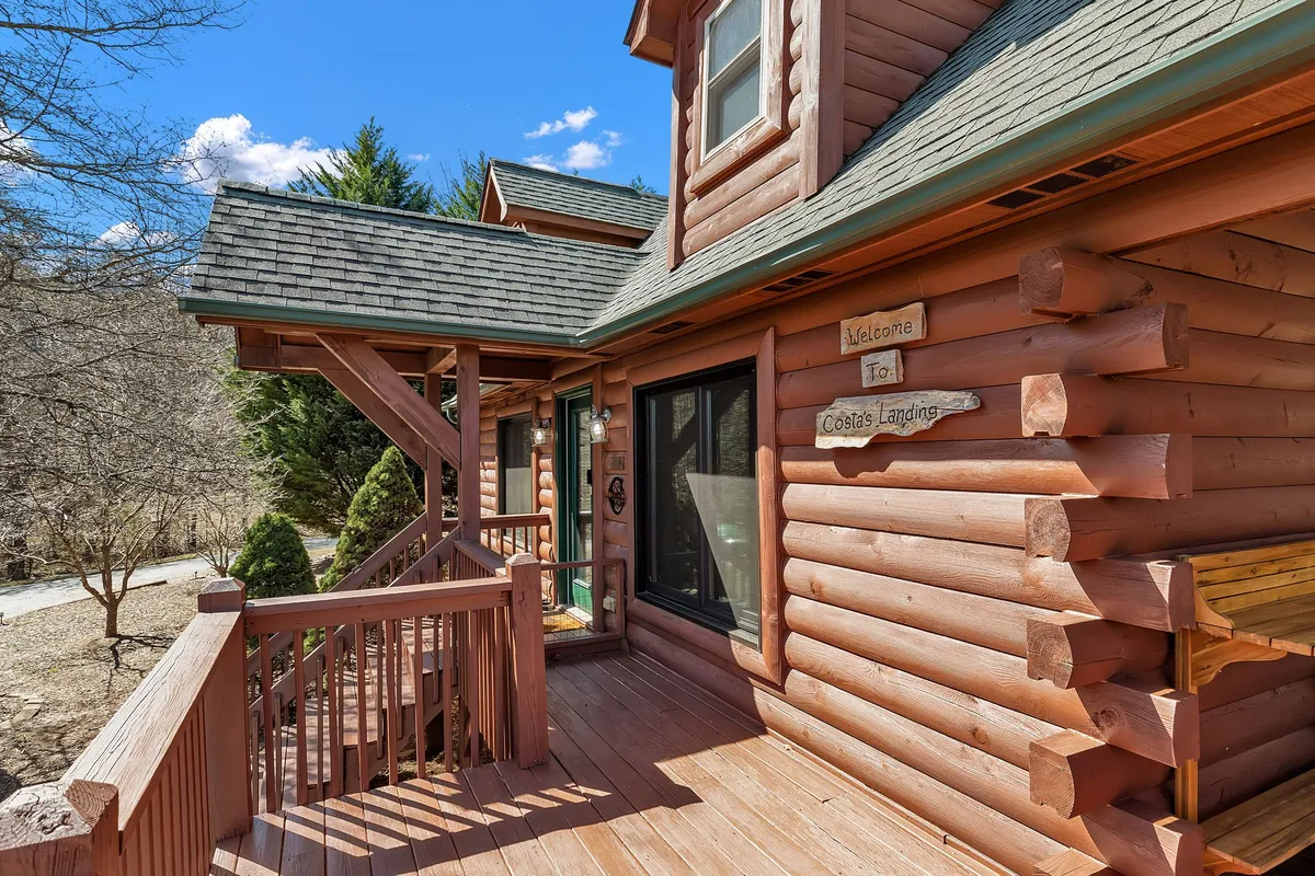 A charming exterior shot showcasing distinctive log cabin architecture, wooden railings, and a welcoming sign reading 'Welcome to Coslas Landing.' The cabin is nestled among greenery, highlighting the enclave-like setting and sense of belonging.