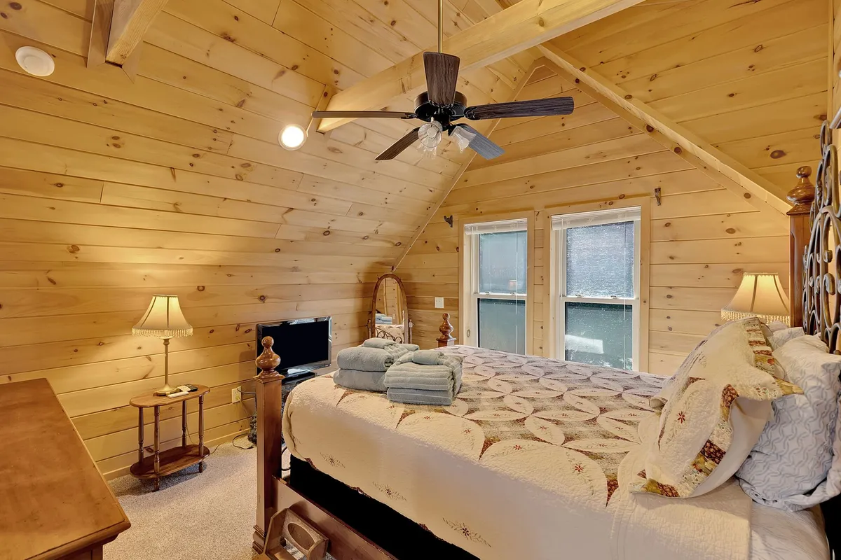 A warm and welcoming bedroom featuring a bed with a decorative quilt, wooden ceiling, nightstands with lamps, and a television. Natural light streams in, highlighting the cozy and inviting atmosphere.