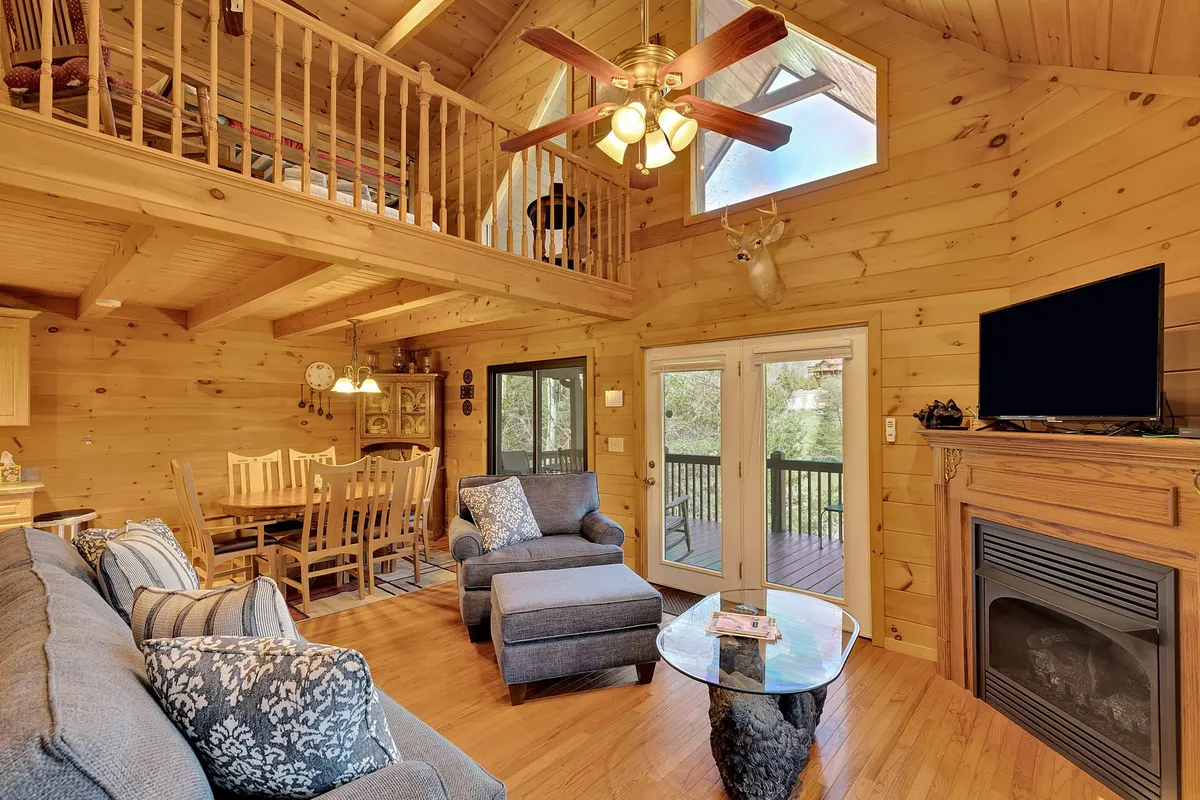 A spacious main living area featuring a cozy fireplace, comfortable seating, and large windows that let in abundant natural light. The room’s wooden ceiling with exposed beams adds warmth, and the arrangement invites gatherings for conversation, reading, or family time. The windows offer glimpses of the outdoor deck and lake beyond.