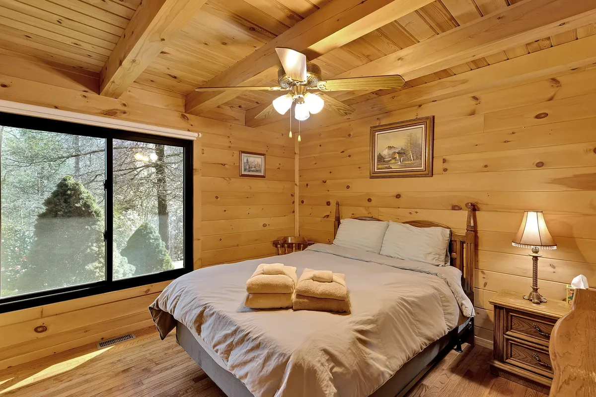 A tranquil bedroom scene with a comfortable bed positioned near a window. Soft, natural light filters in, illuminating the nightstand and highlighting the peaceful setting ideal for quiet mornings, sipping coffee, or unwinding together while enjoying nature views.