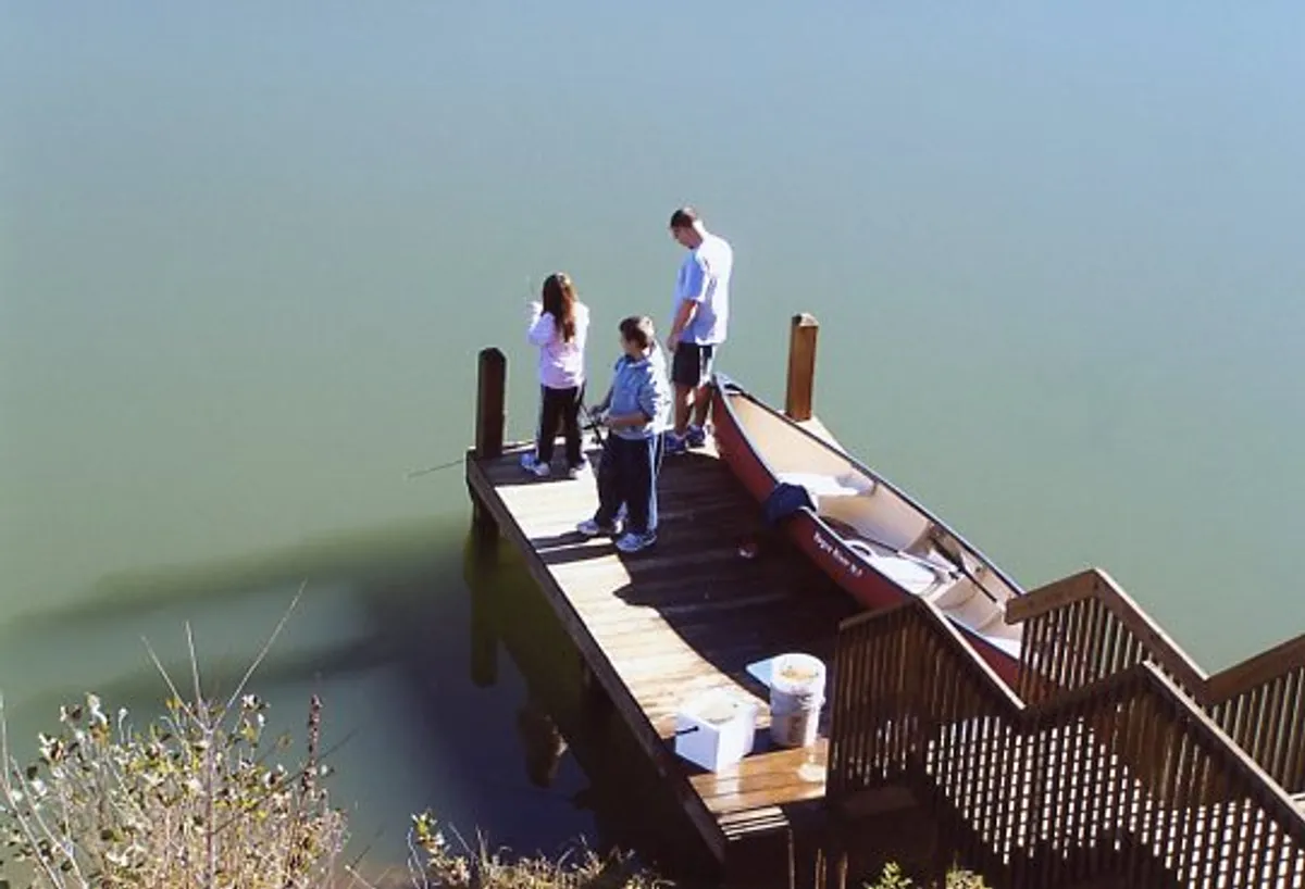 A lively wooden dock stretches over the lake, with a canoe tied up alongside. Children are seen fishing off the end while an adult supervises, all beneath a glowing sunset sky. The water is calm, and everyone appears relaxed and joyful.