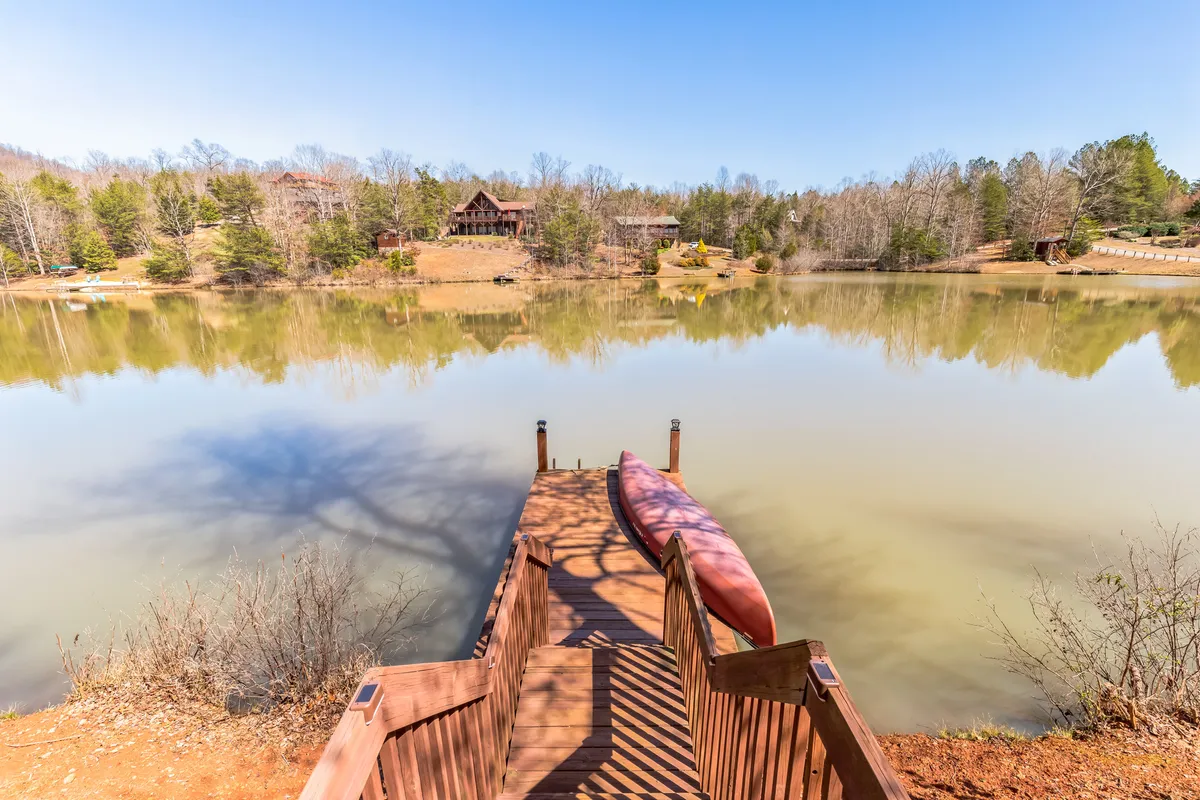 A peaceful lakeside scene features a wooden dock with a kayak, surrounded by calm water and trees, with a distant cabin visible. The area looks ideal for relaxing, wildlife watching, or enjoying a campfire by the shore.
