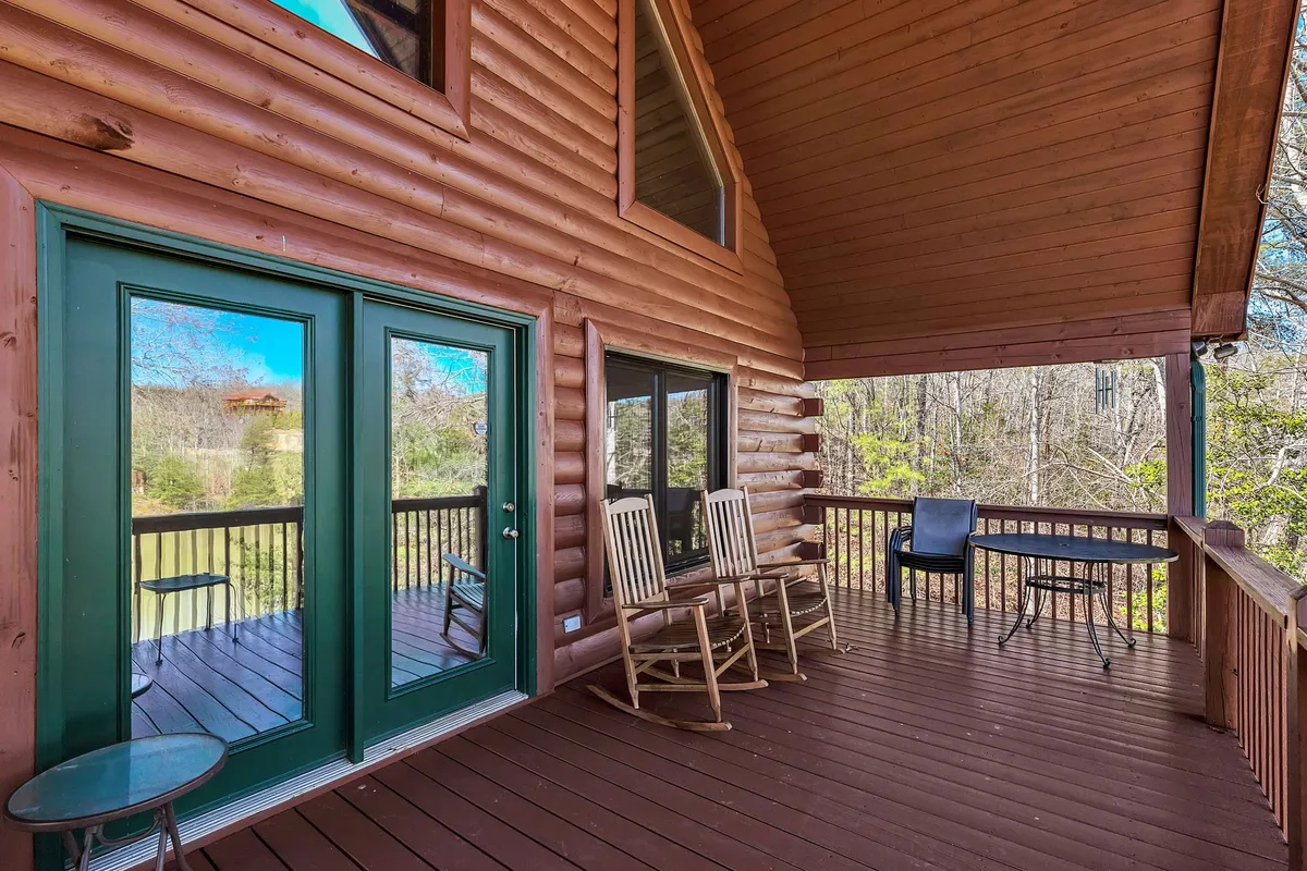 The inviting exterior of the cabin features green double doors, classic rocking chairs on a wooden deck, and the lush backdrop of surrounding nature. The scene evokes early mornings with coffee or peaceful evenings soaking in the view and socializing.