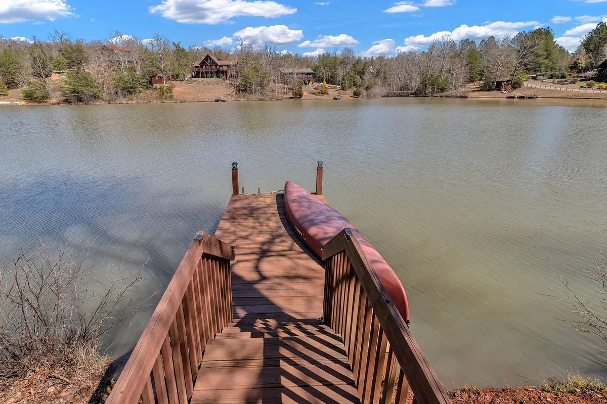 A wooden dock stretches out onto the tranquil waters of Laurel Lake, with a canoe ready to launch. The dock is surrounded by lush trees, and a few distant houses can be seen dotting the shoreline. The scene is calm and inviting, perfect for boating, fishing, and enjoying family time by the water.