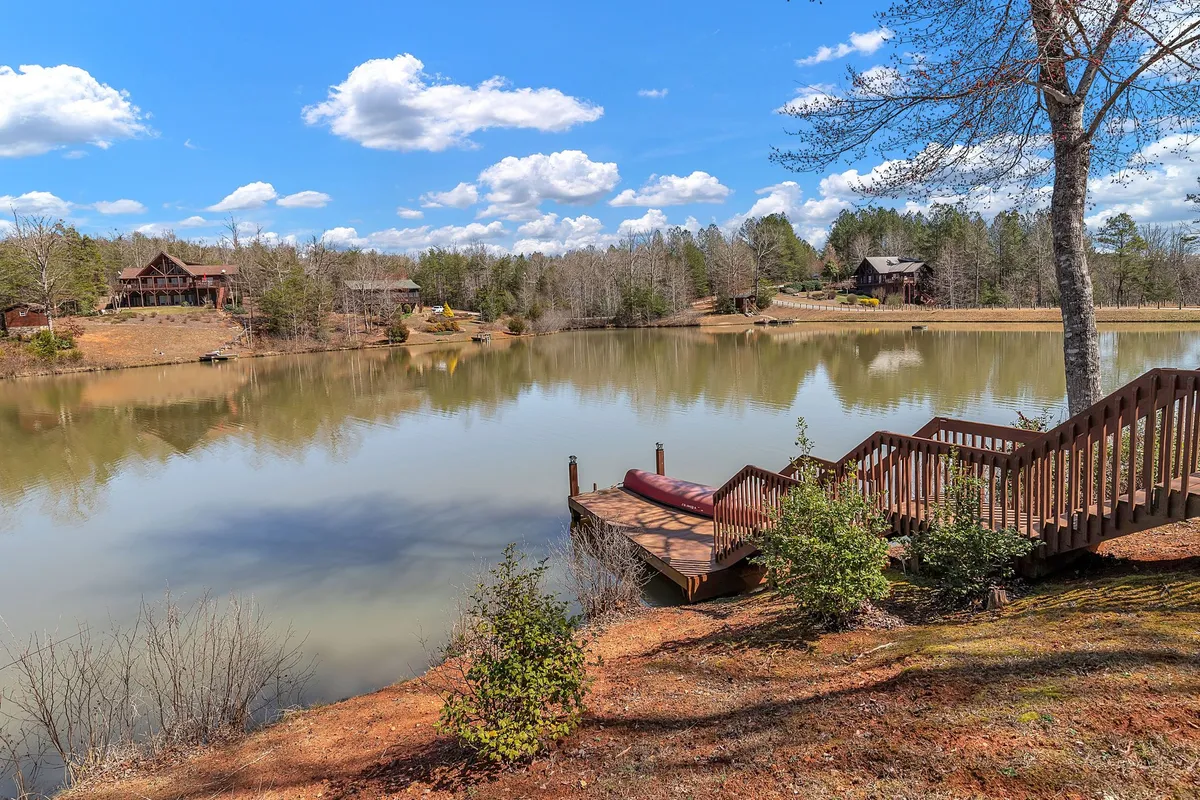A serene view of a calm lake, bordered by a wooden dock and lush surrounding trees with distant cabins visible across the water. The image conveys peacefulness, privacy, and access to nature, while the absence of motorboats reinforces the quiet, safe environment.